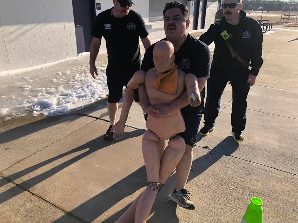 Man carrying human dummy for firefighter training with two men following him