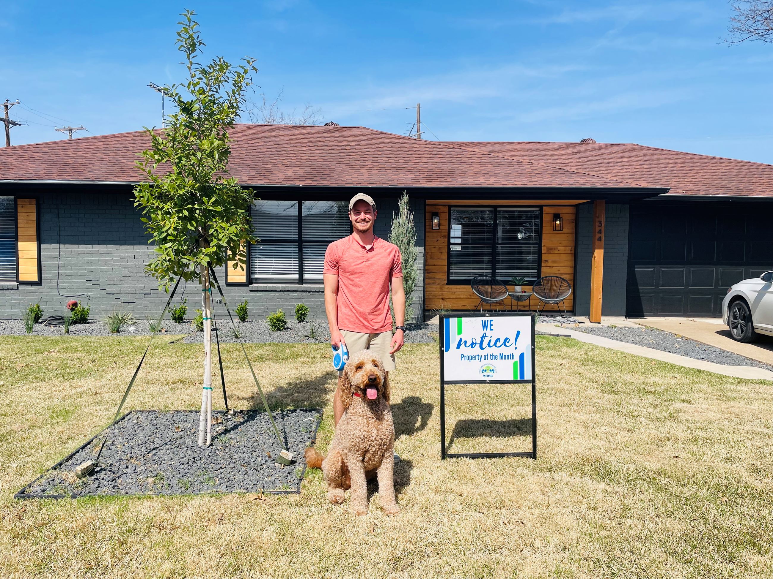 Man with dog standing in front of a black house with a "Property of the Month" yard sign